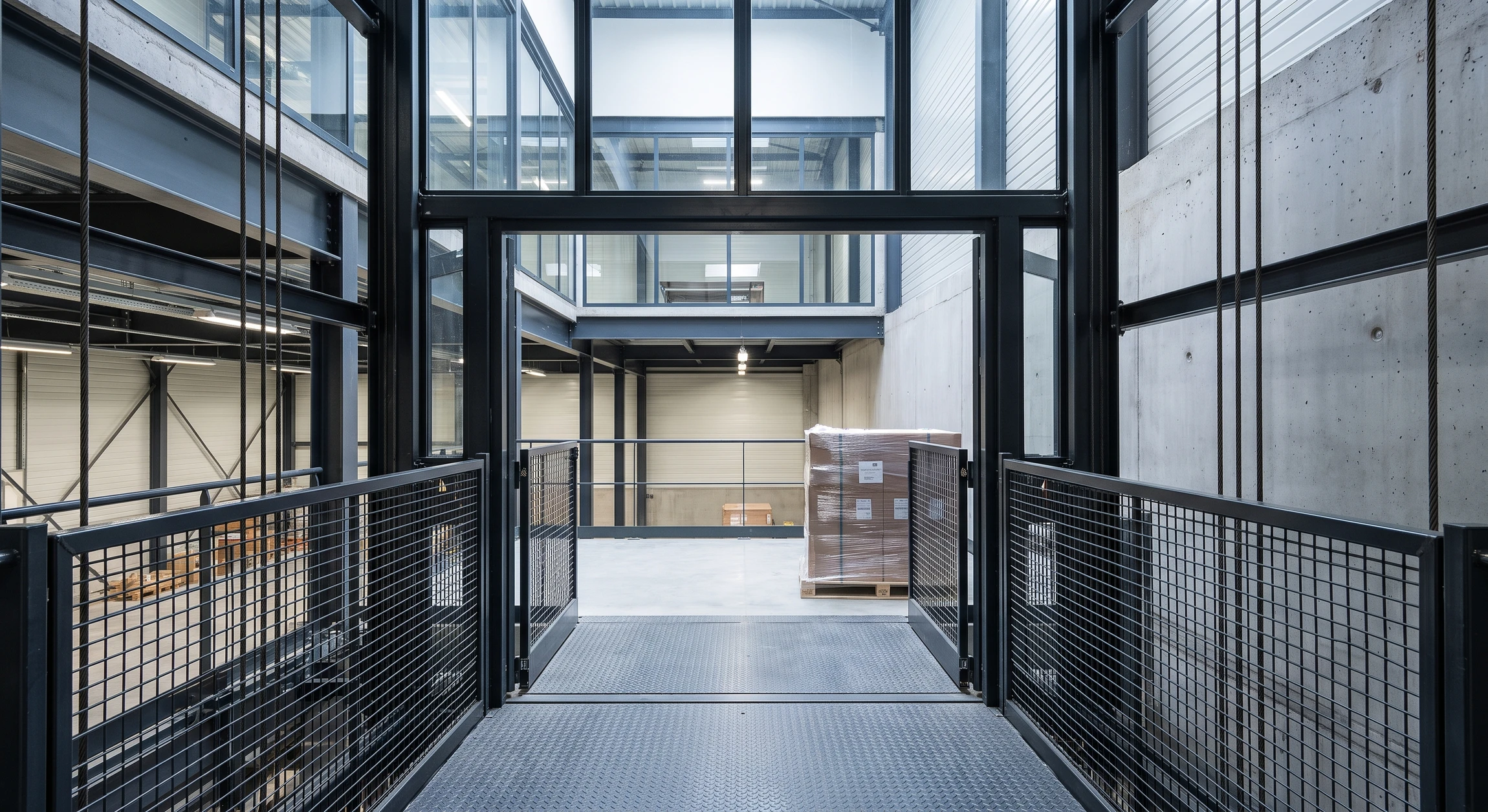 Photographie réaliste d'un monte-charge industriel moderne installé dans une cage d'escalier ou une ouverture de bâtiment, capturant la structure métallique robuste, les câbles de suspension et la cabine de transport, dans un environnement de chantier ou de bâtiment commercial modéré. La mise en scène doit montrer l'équipement comme un système d'élévation vertical professionnel, avec une perspective qui souligne sa capacité à optimiser les flux verticaux de charges étage après étage.