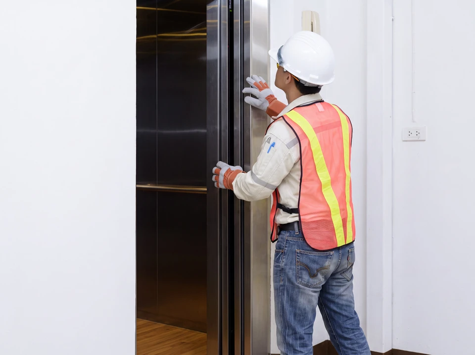 Technicien ascensoriste en uniforme professionnel effectuant une inspection/maintenance préventive d'un ascenseur ou d'une porte automatique dans un immeuble (ERP). La scène montre le professionnel contrôlant les mécanismes de l'équipement ou les portes, avec outils professionnels visibles, illustrant concrètement l'action de maintenance préventive régulière mentionnée dans le contenu de la section.