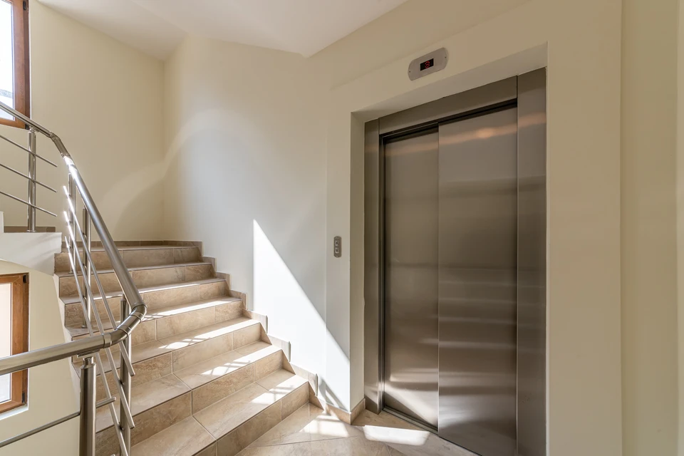 Deux ascenseurs modernes dans un hall d'immeuble contemporain, l'un ouvert révélant l'habitacle éclairé avec main courante et parois inox, l'autre fermé avec portes coulissantes métalliques