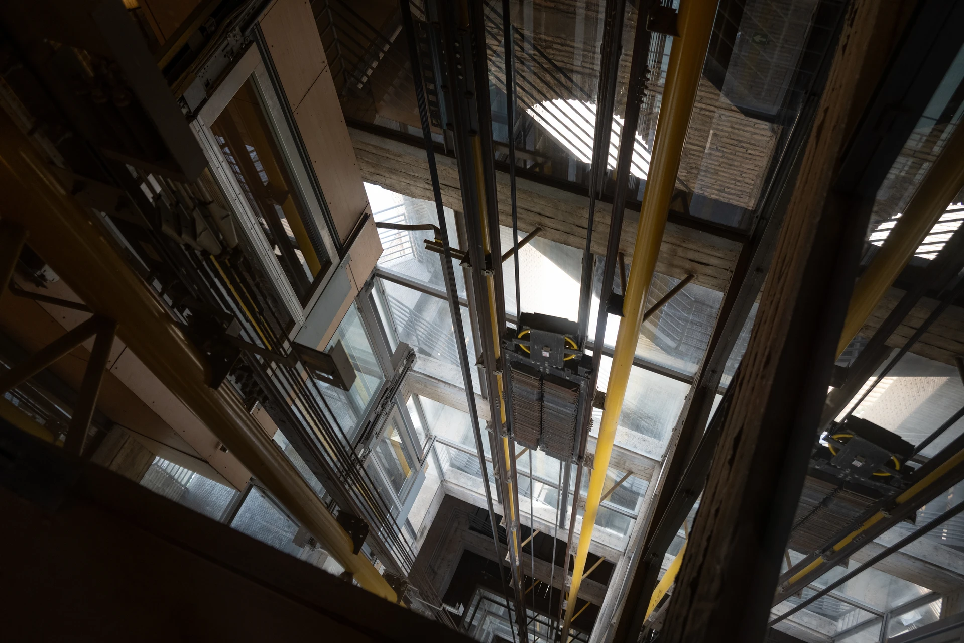 Photographie en couleur d'une déconstruction d'ascenseur en cours dans une cage d'escalier ou une gaine d'immeuble. La scène montre des techniciens en équipements de sécurité (casques, gilets de protection, gants) procédant au démontage méthodique d'une cabine d'ascenseur ou de ses composants (rails, poulies, câbles). L'environnement de travail est professionnel avec des mesures de sécurité visibles (barricades, zones délimitées). La photo doit transmettre rigueur, professionnalisme et respect des normes de sécurité inhérentes aux opérations de déconstruction spécialisée en milieu urbain.