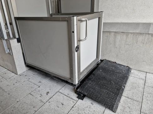 Intérieur d'une cabine d'ascenseur en cours de rénovation esthétique, montrant un technicien en tenue professionnelle installant des panneaux de revêtement moderne sur les parois intérieures de la cabine. La cabine affiche un contraste net entre les parois anciennes (gris neutre) et les nouvelles surfaces en cours d'installation (matériau contemporain type métallique ou composite), avec outils de rénovation visibles. L'éclairage professionnel et les détails techniques renforcent l'aspect de travail spécialisé en matière d'habillage de cabine.